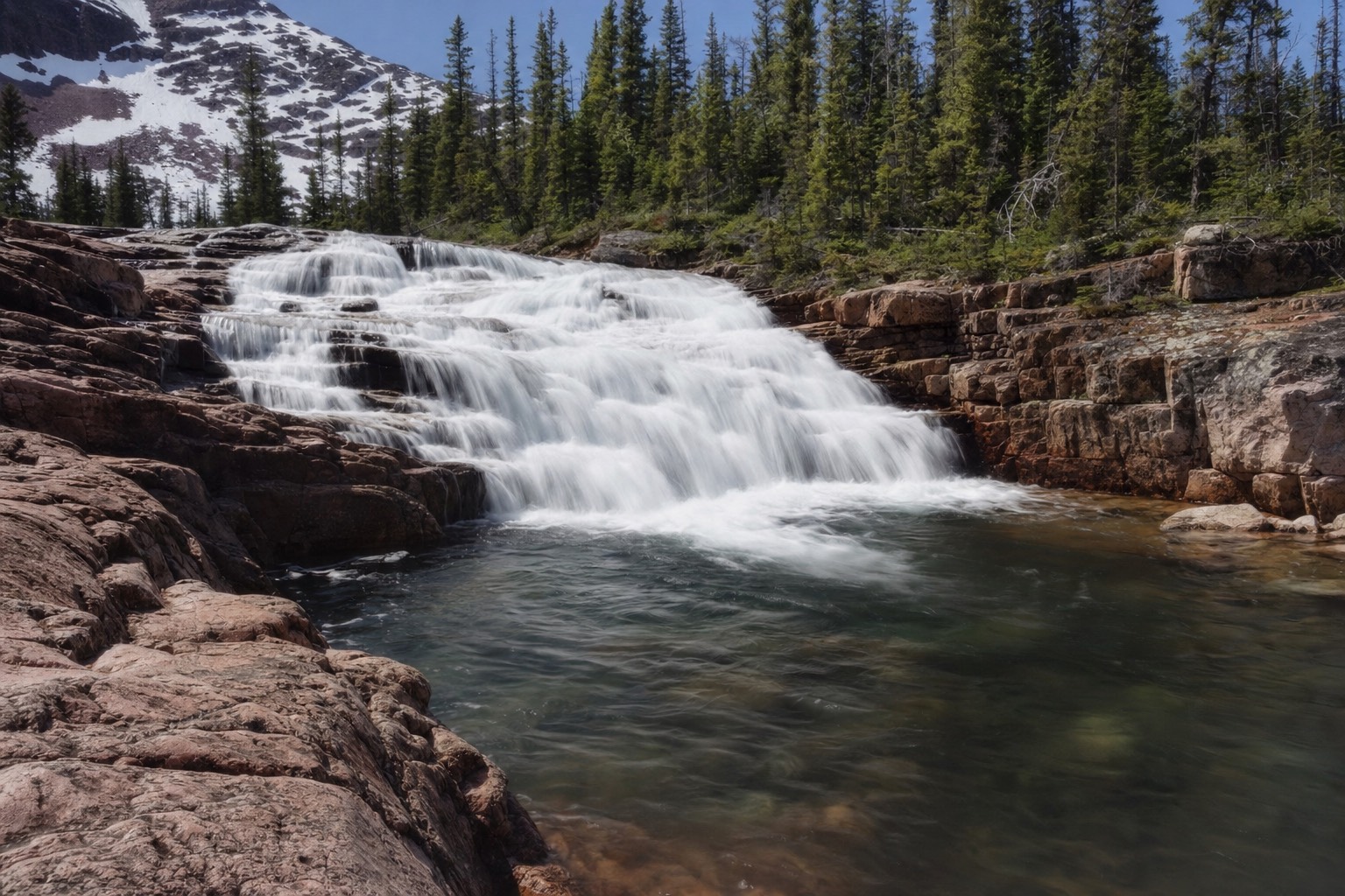 Cameron Falls pouring over pink granite ledges into a pool ringed by spruce on a bright northern summer day