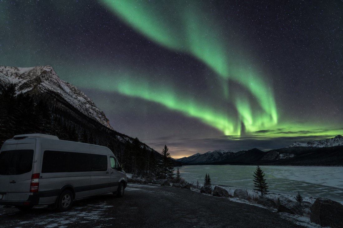 Green aurora bands over a dark frozen lake beside the Ingraham Trail with a parked tour van in the foreground