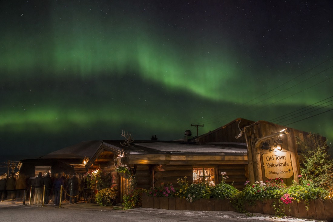 Low log cabin restaurant with a sign and flowers in summer, diners waiting outside in Old Town Yellowknife