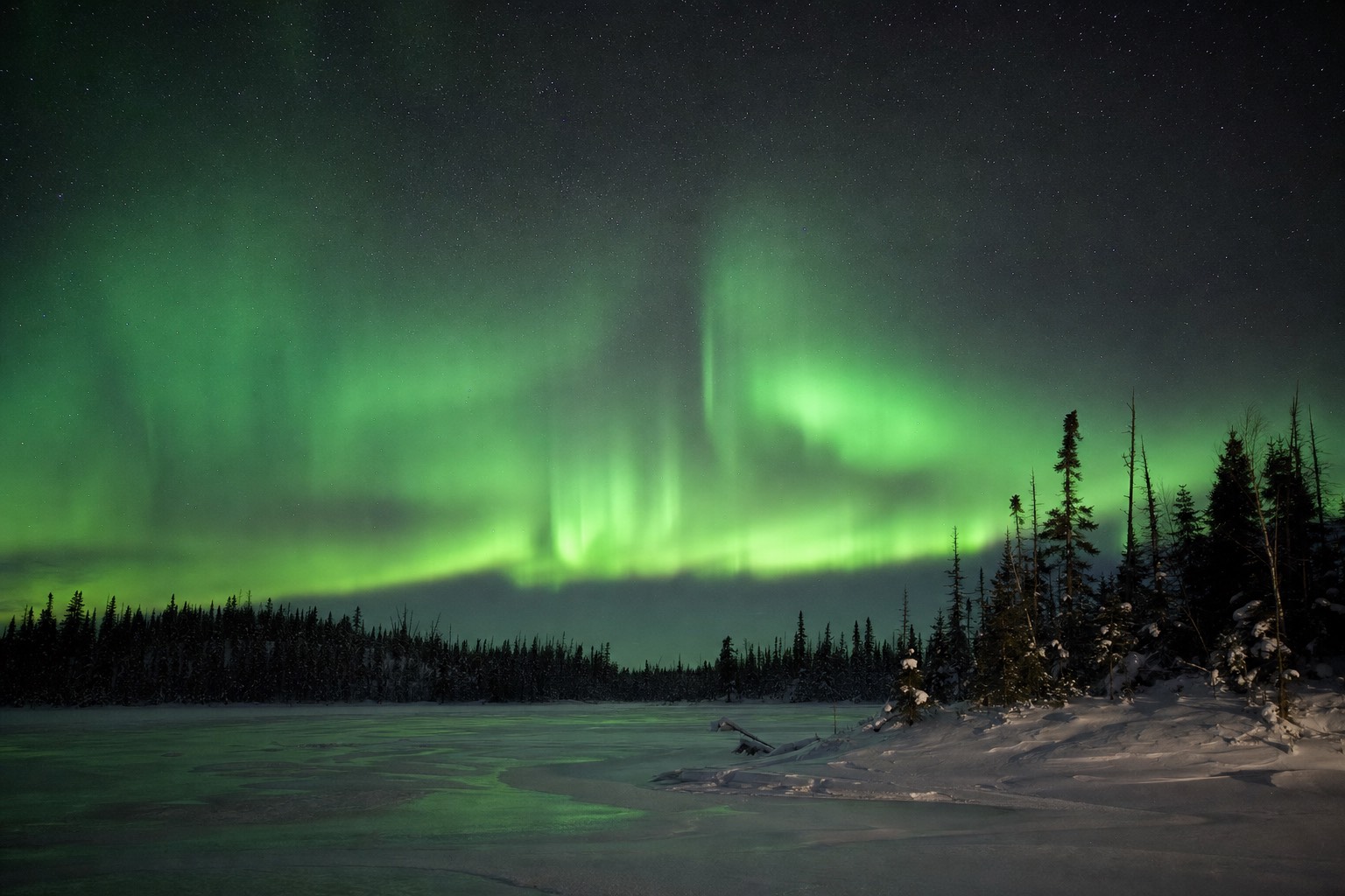 Green aurora curtains overhead on a frozen northern lake, far brighter than a mid-latitude horizon glow