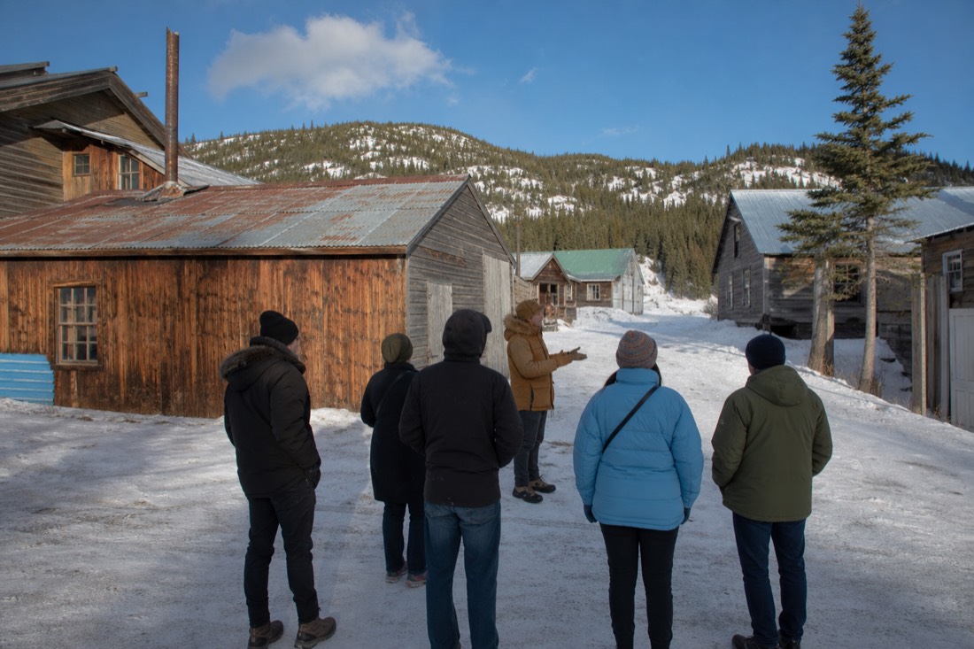 Small tour group listening to a guide beside weathered gold-rush shacks on a bright winter afternoon in Old Town