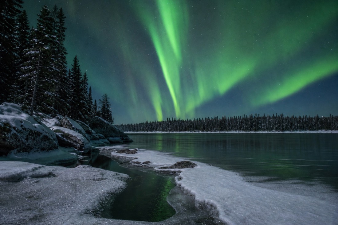 Pale aurora arc strengthening into green curtains over a snow-covered lake at midnight