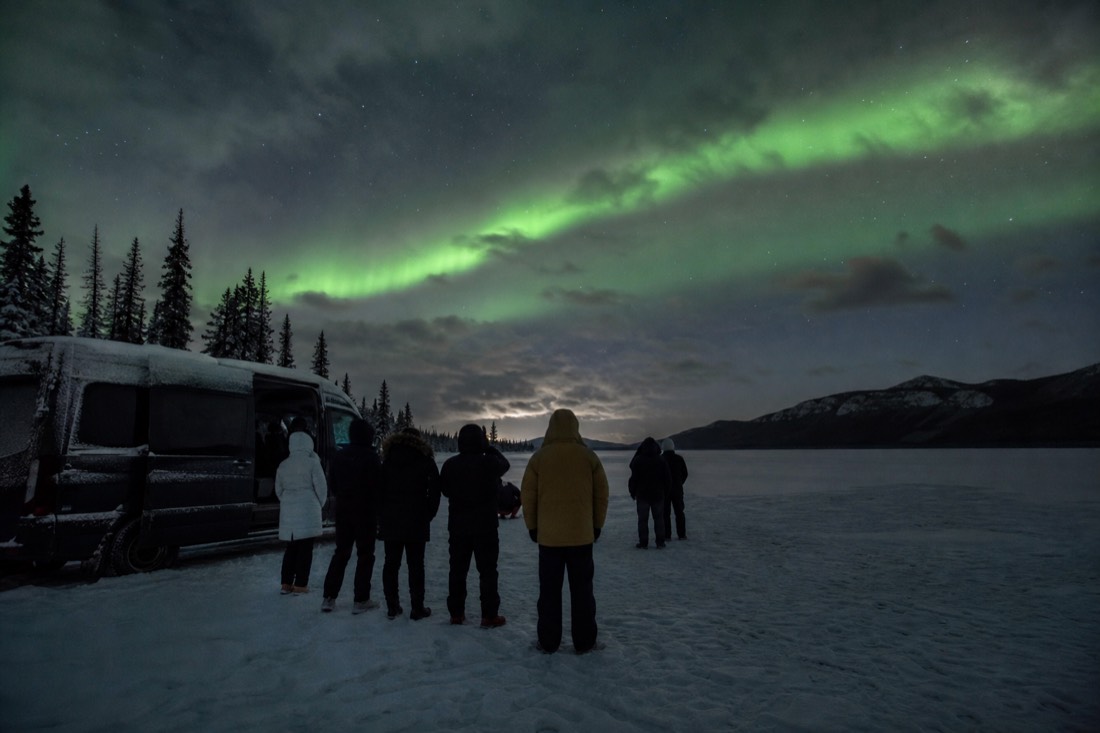 Guests waiting under an overcast night sky beside a tour van on a frozen lake
