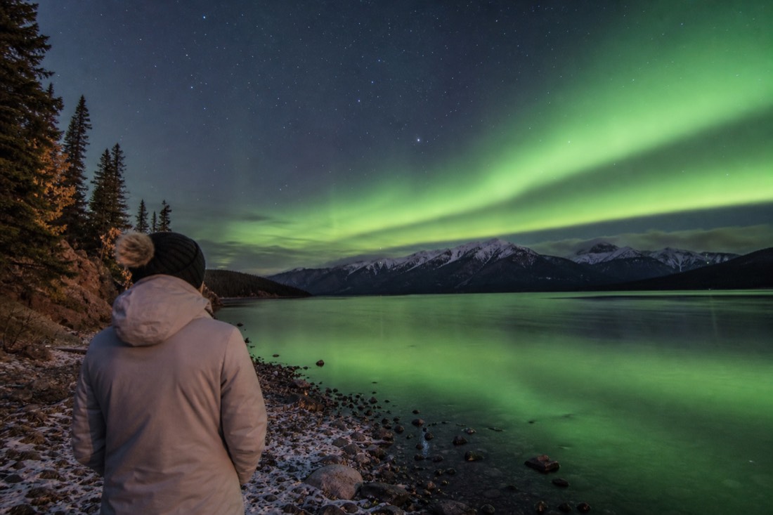 Traveler in a light jacket and toque watching the night sky from a lakeshore in autumn