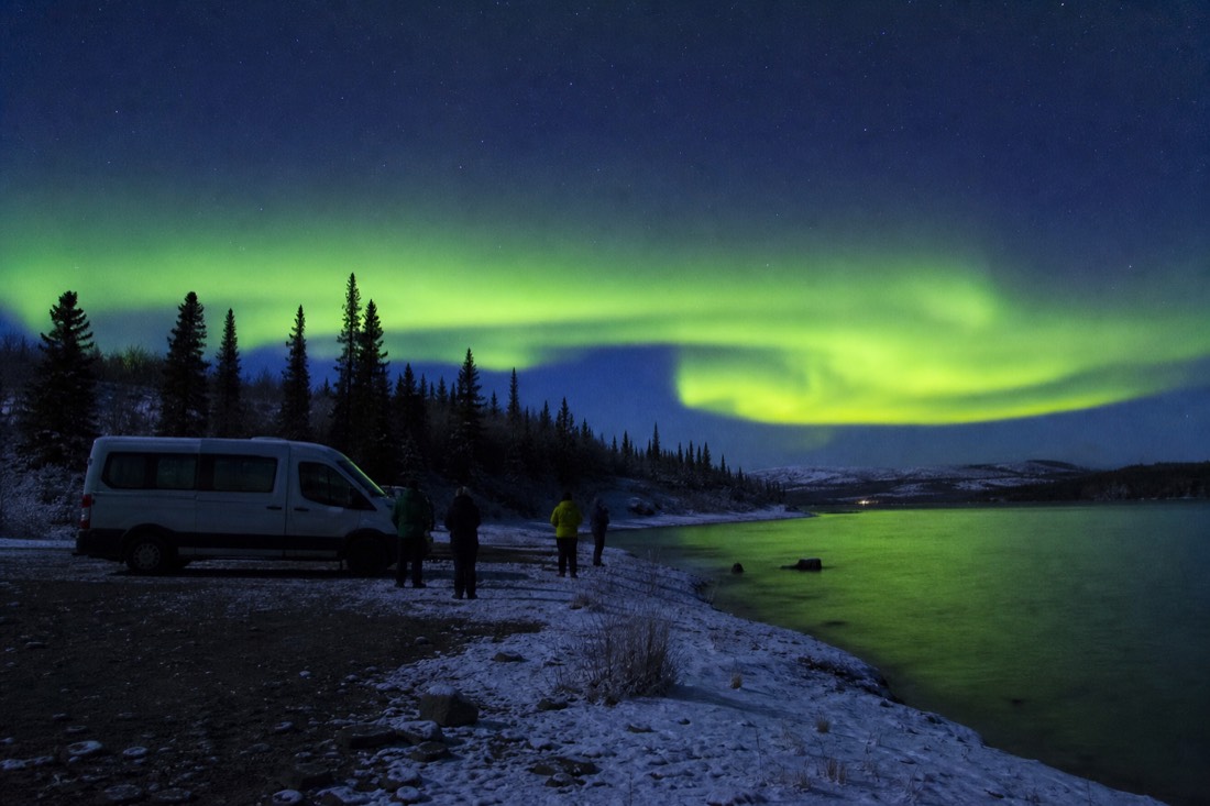 Small group of aurora watchers beside a tour van on a quiet gravel lakeshore at night