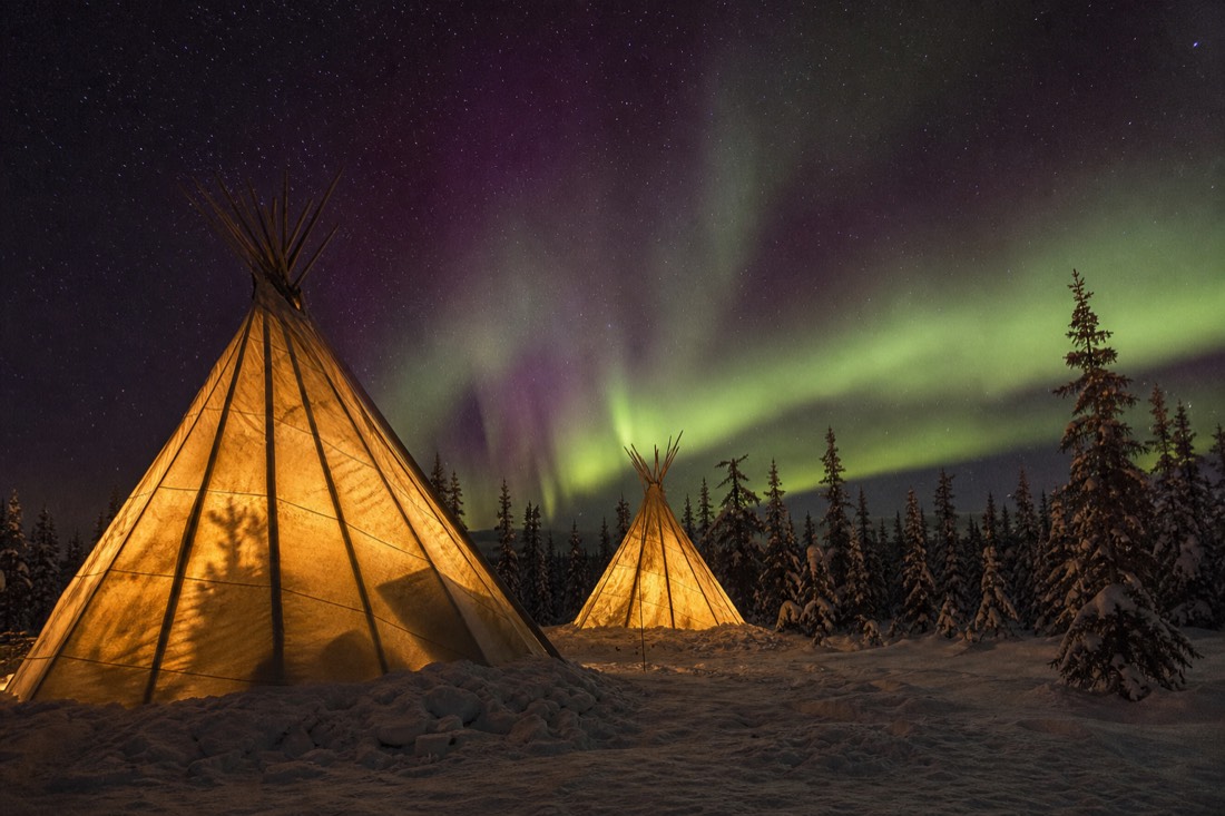 Glowing illuminated teepees in a snowy forest clearing under a starry northern sky