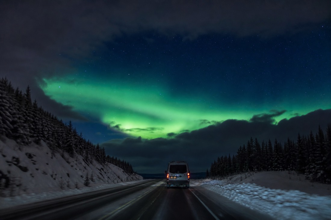 Van driving a dark winter highway toward a band of clear starry sky beyond a cloud edge