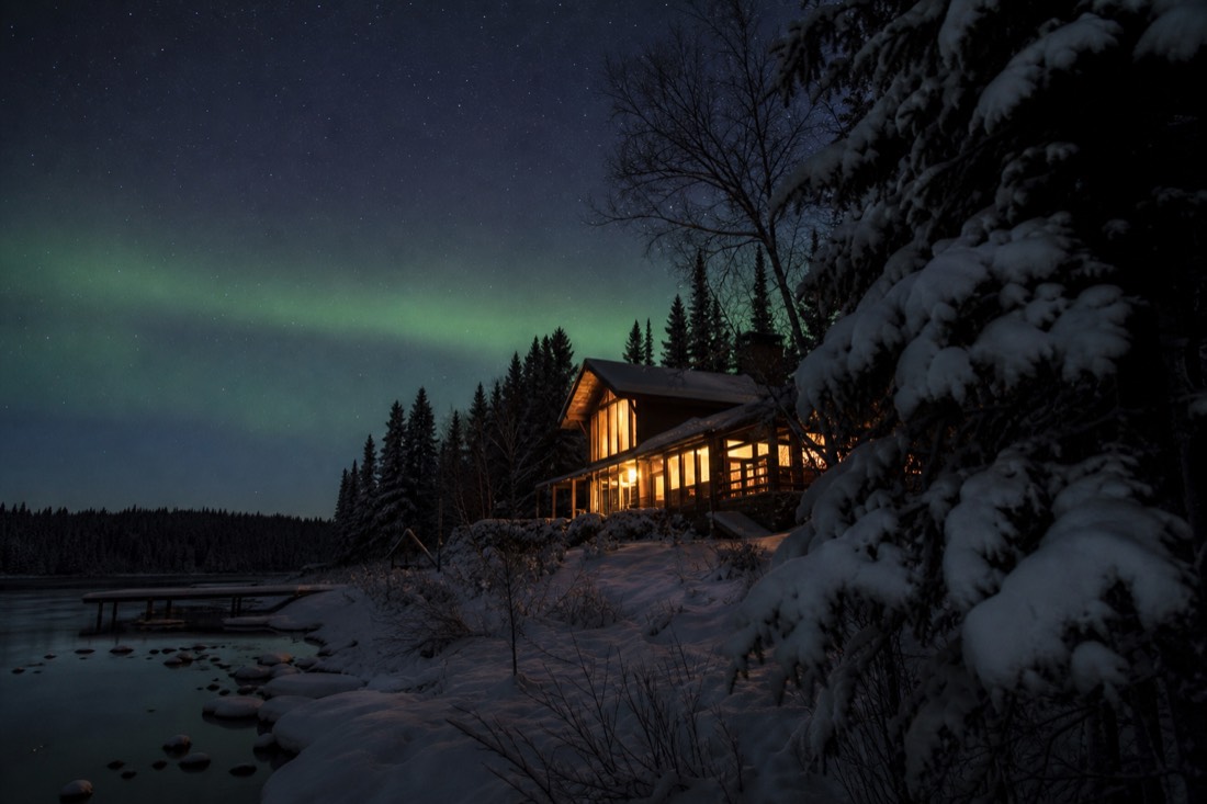Warm lit lakeside lodge windows glowing under a faint green aurora in a snowy clearing