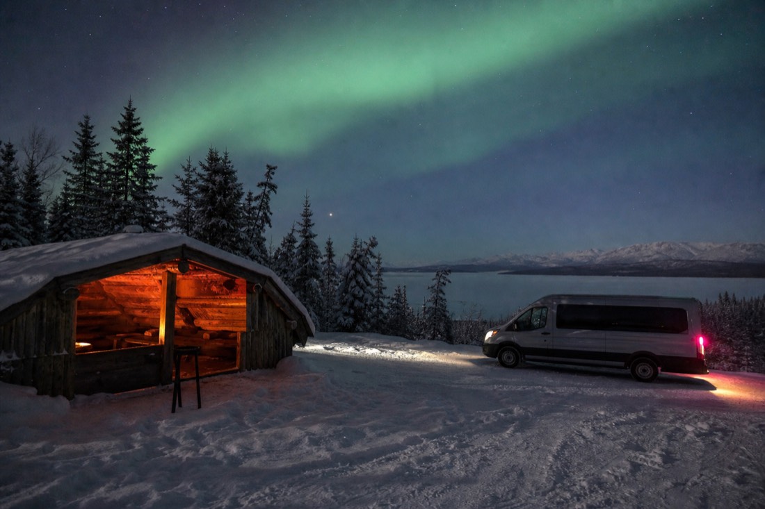 Snowy lakeside pullout with a heated shelter on one side and a tour van idling on the other under faint green aurora
