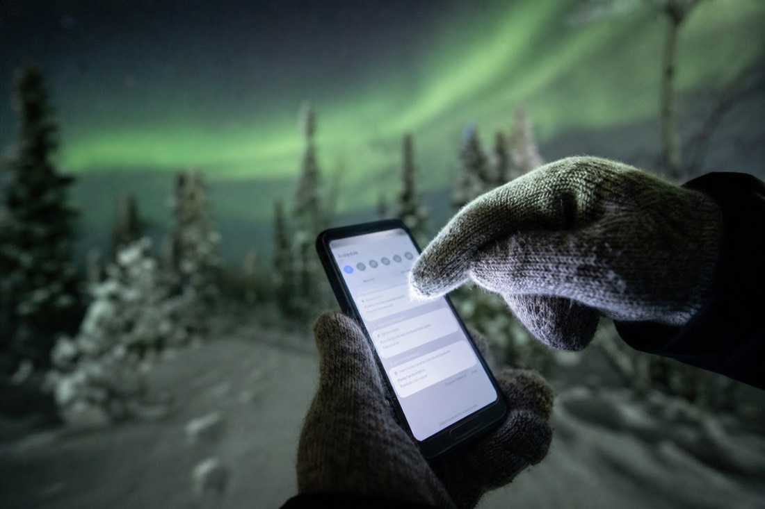 Close-up of a mittened hand tapping a phone notification screen with a snowy treeline and green sky behind