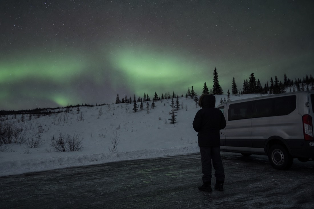 Traveller in a heavy parka standing beside a tour van on a dark roadside pullout under faint green aurora