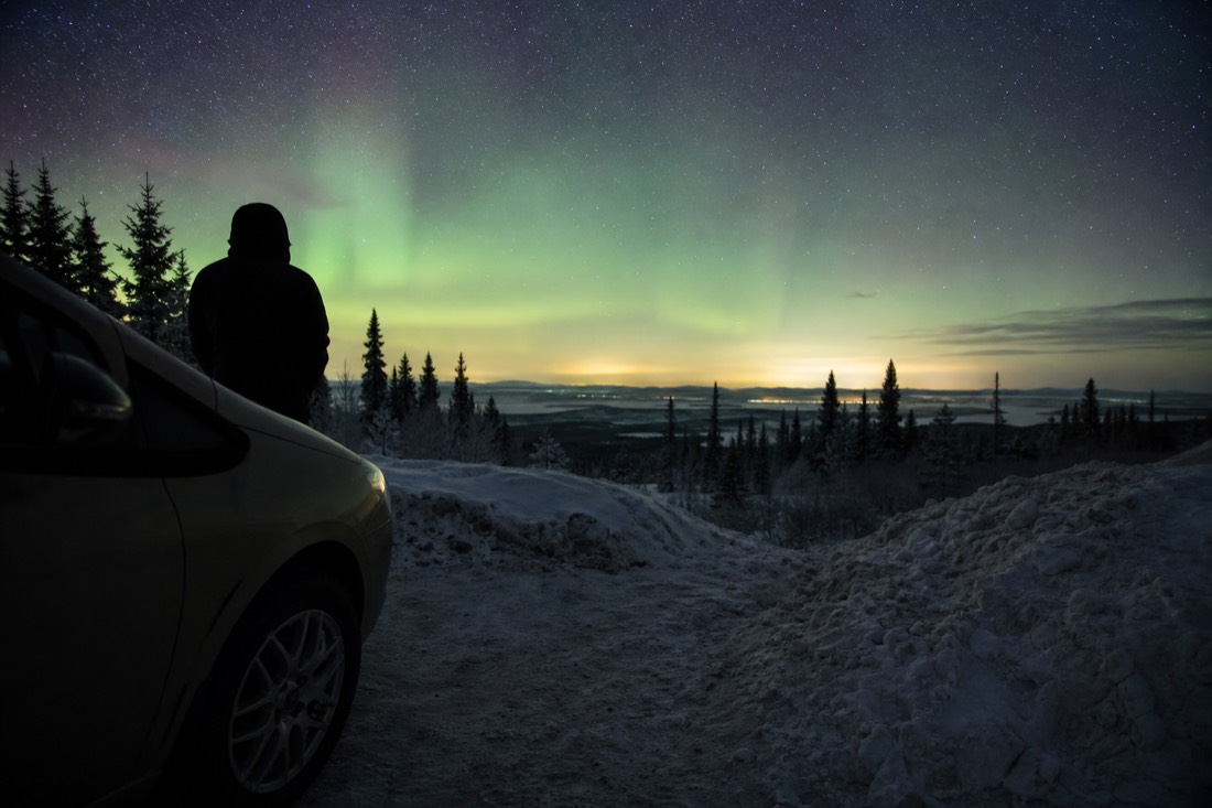 Traveler checking the night sky from a dark roadside viewpoint with a faint green glow on the horizon