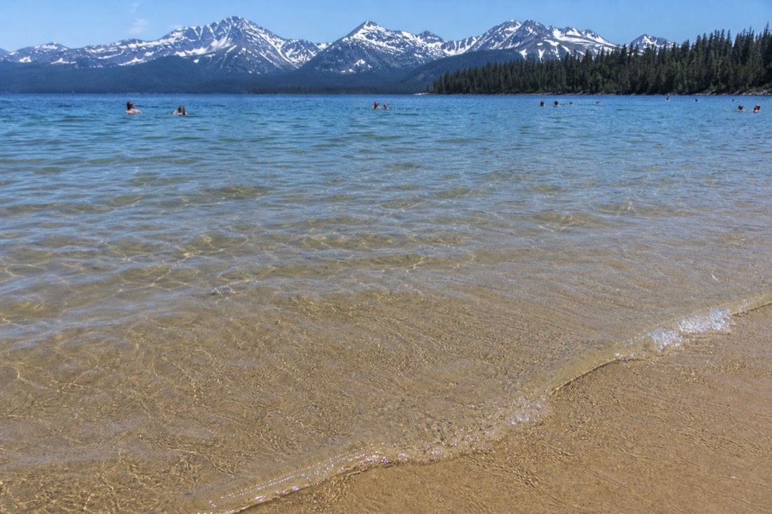 Clear shallow lake water over sand at a northern beach with swimmers further out on a summer afternoon