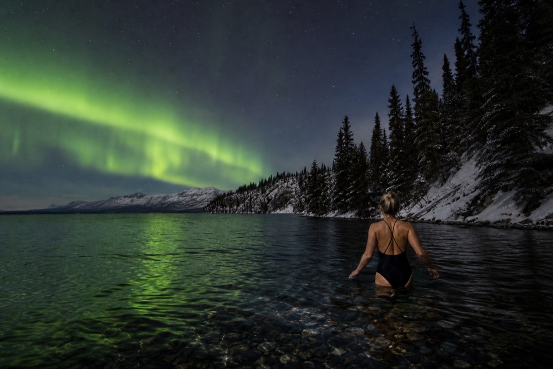 Swimmer wading into bright cold lake water with goosebumps visible and a rocky point in the distance