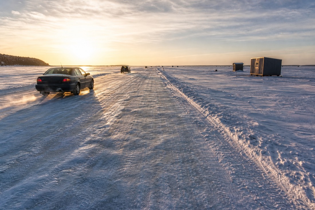 Cars driving a plowed ice road across the frozen bay past fishing huts under low winter sun