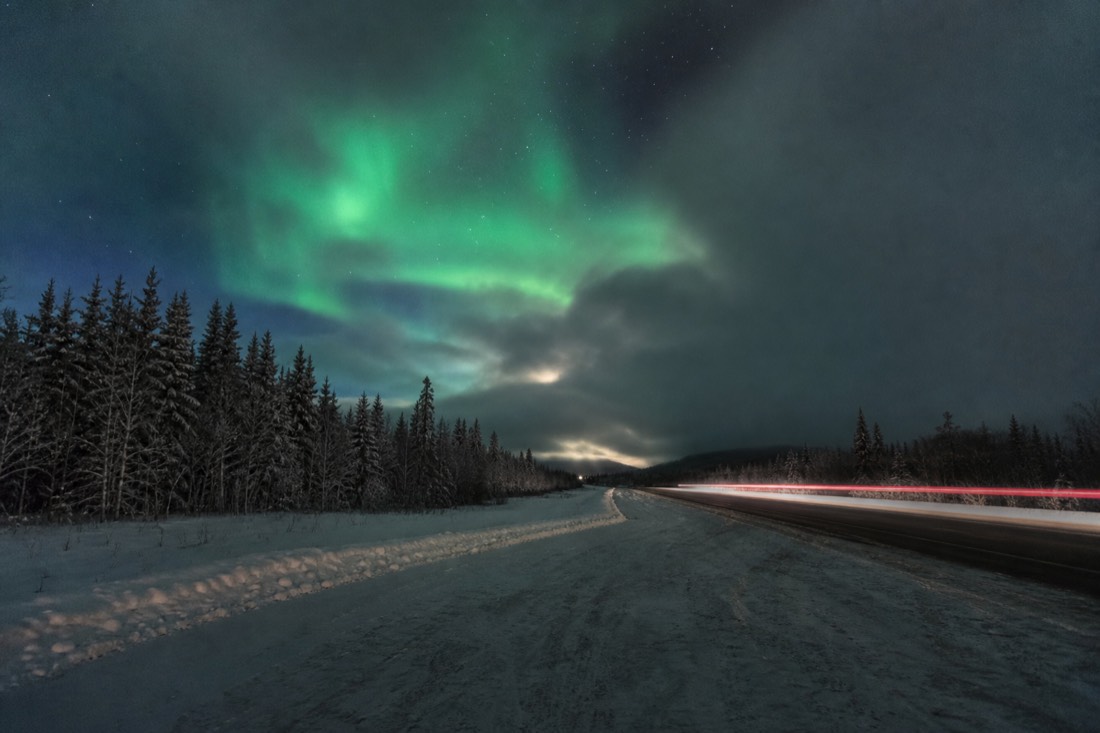 Aurora glowing through a break in fast-moving clouds over a highway pullout at night