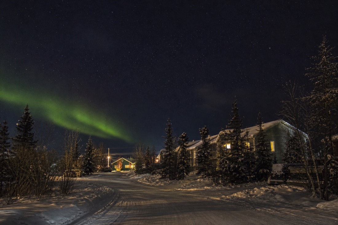 Faint green aurora visible above rooftops from the edge of a quiet Yellowknife residential street at night