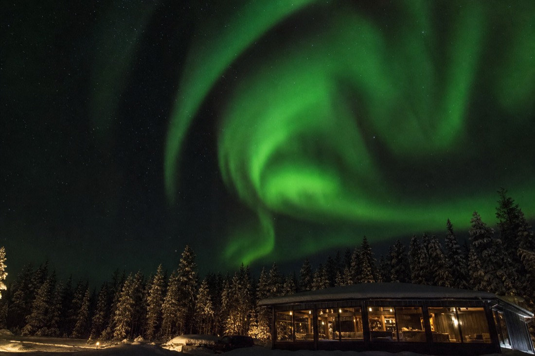 Brilliant green aurora spirals filling a fully dark winter night sky above a heated viewing cabin