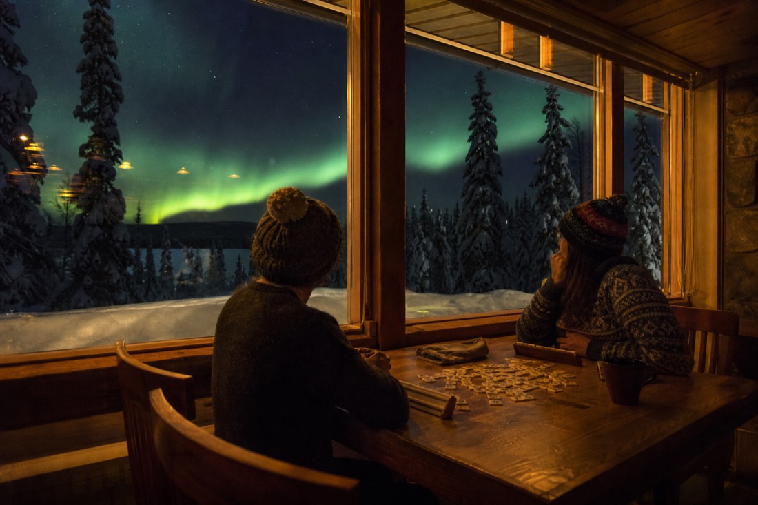 Warmly lit lodge interior with guests playing games by the window while snow and darkness sit outside