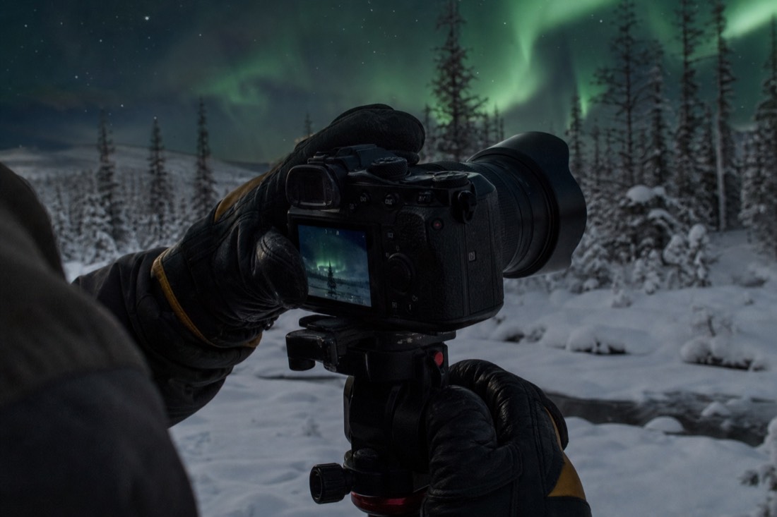 Close view of gloved hands adjusting a camera on a tripod under a green-lit night sky