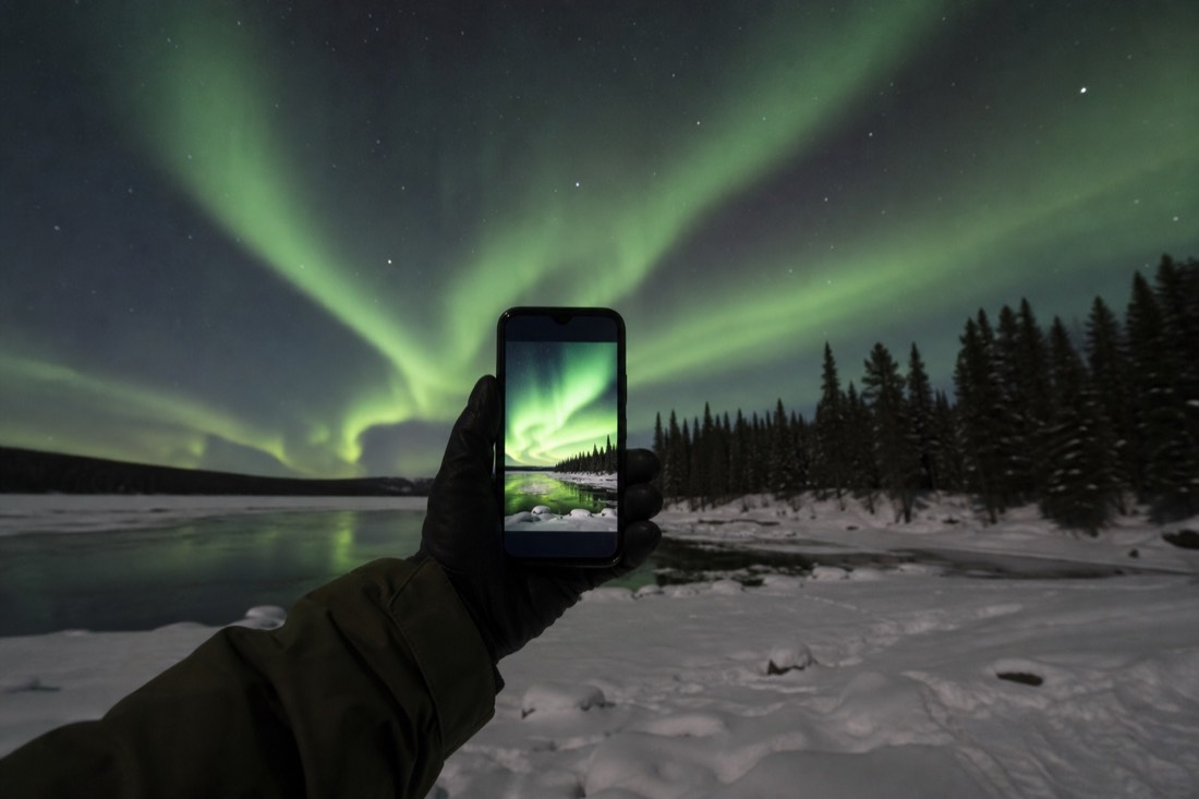 Traveler holding up a phone showing a vivid aurora photo with the real aurora behind it over a frozen lake