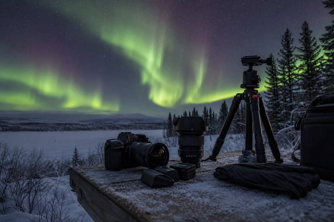 Aurora photography kit laid out on a wooden table with tripod, wide lens, spare batteries, and thin gloves