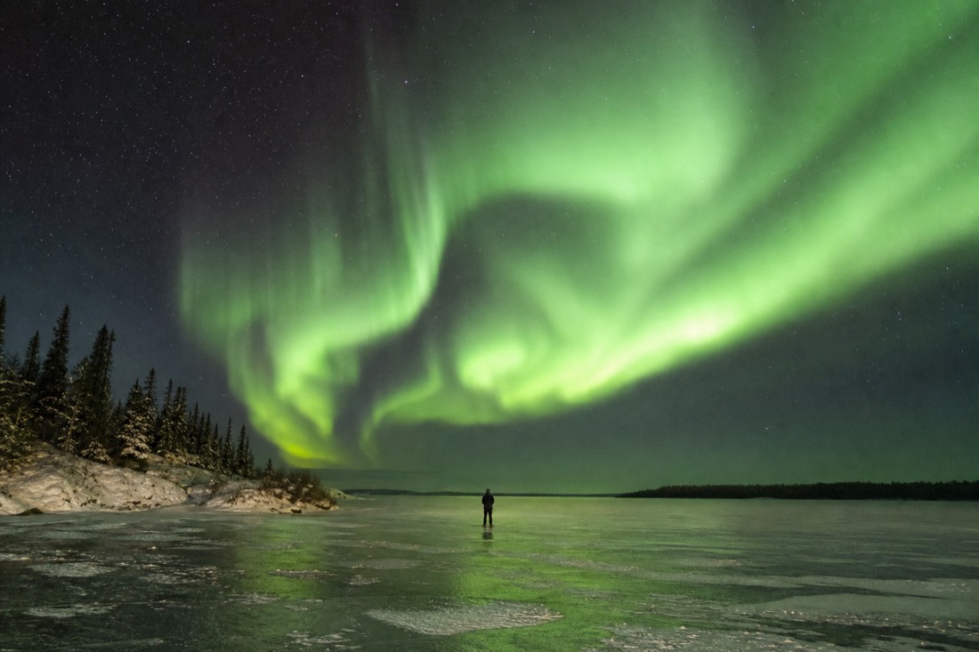 Green aurora filling the sky above a low flat lake horizon with a single small figure standing on the ice