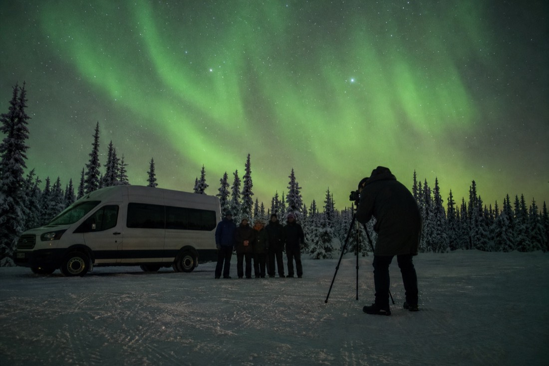 Tour photographer positioning guests for a portrait under bright aurora beside a parked van