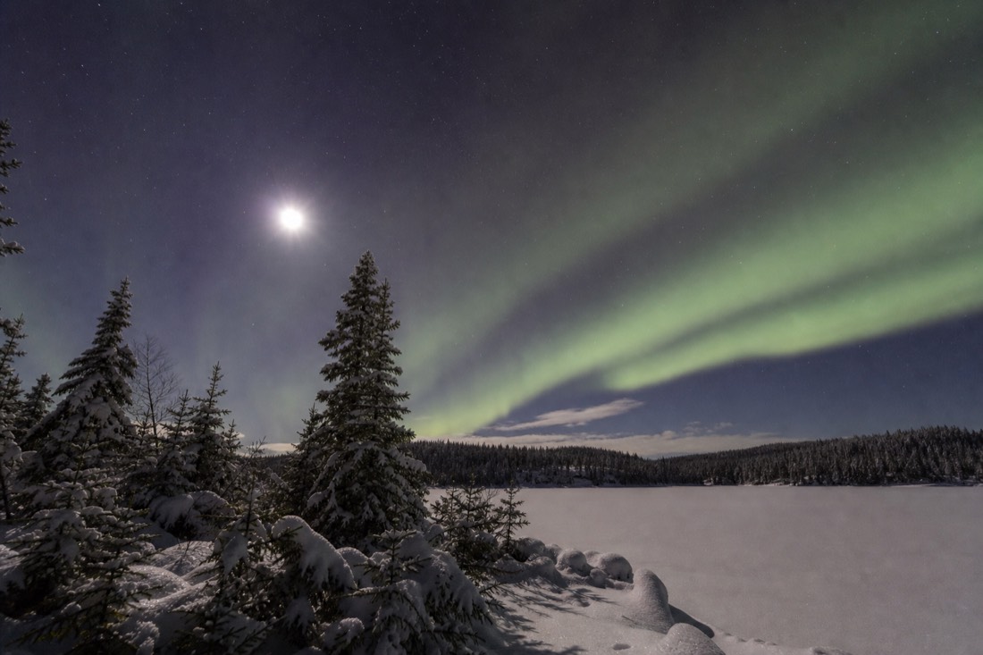 Faint grey-green aurora band over a moonlit snowy lake with stars visible around it