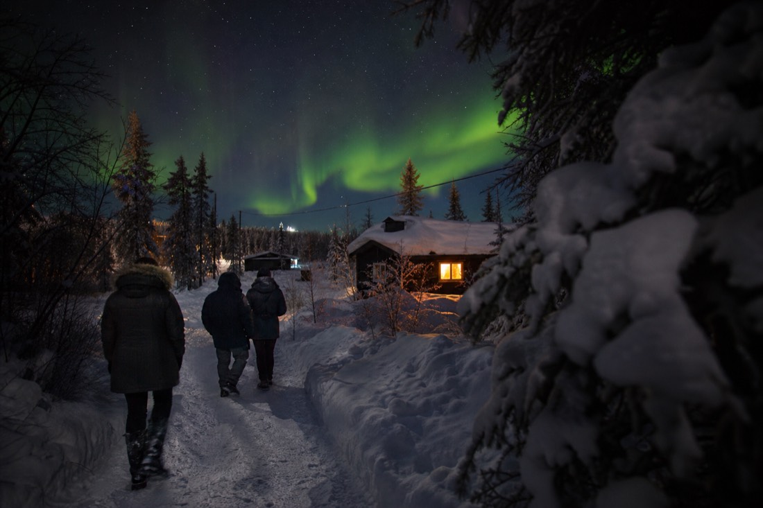 Visitors walking a snowy Old Town Yellowknife lane in low afternoon light with a warm lit cabin window ahead