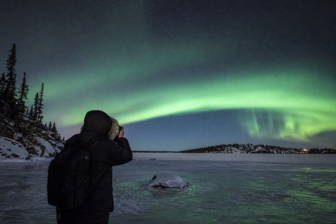 Traveler in a heavy parka photographing a wide green aurora arc over a frozen lake near Yellowknife
