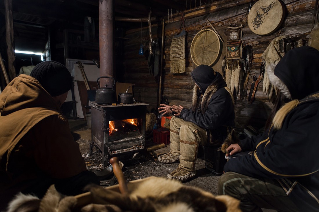Elder telling stories beside a wood stove with drums and beaded hide work on the cabin wall