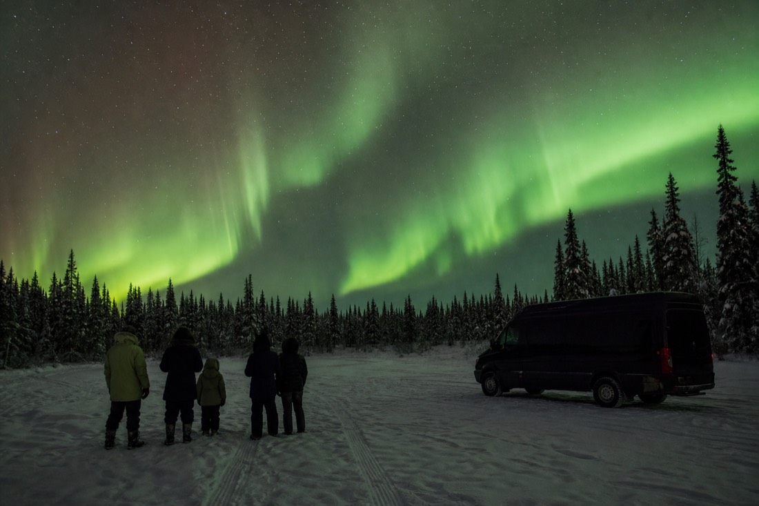Guests in full arctic kit watching green aurora while a heated van waits with its lights off nearby