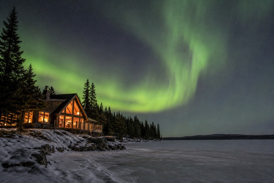 Warmly lit lodge windows glowing beside a frozen lake while green aurora moves across the sky above