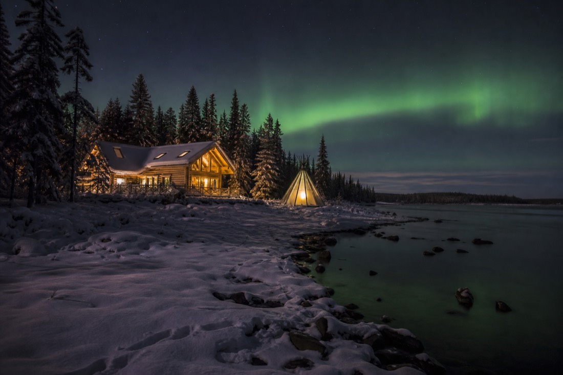 Lit wooden aurora cabin and a glass tipi on a snowy shoreline with a faint green band of northern lights above the treeline