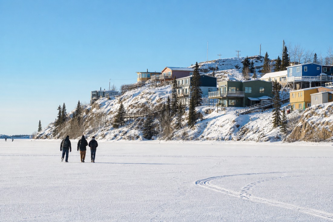People walking on the frozen bay in bright March sunshine with Old Town Yellowknife on the rocks behind