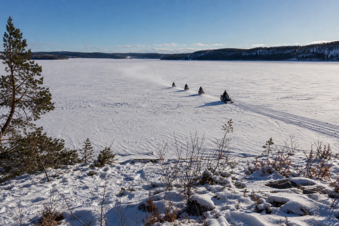 Snowmobiles crossing a wide frozen lake in bright midday March light with long blue shadows
