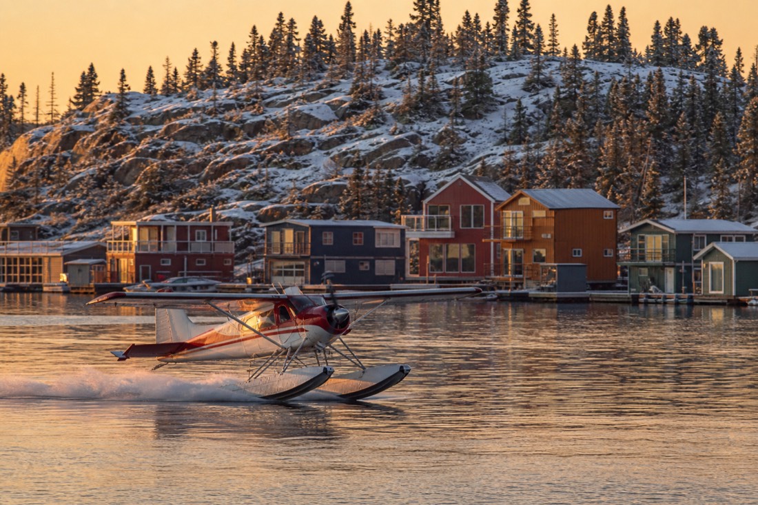 Floatplane lifting off Yellowknife Bay past colourful houseboats in golden evening light