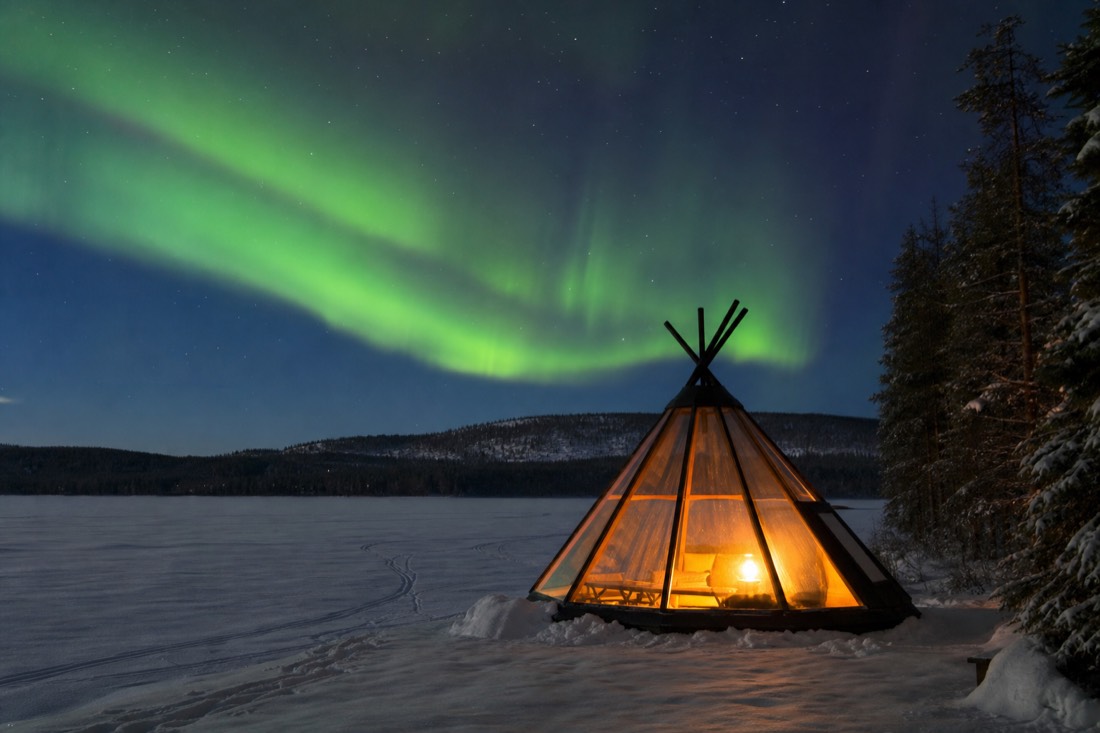 Glass tipi glowing warmly on a frozen lake at night with green northern lights overhead