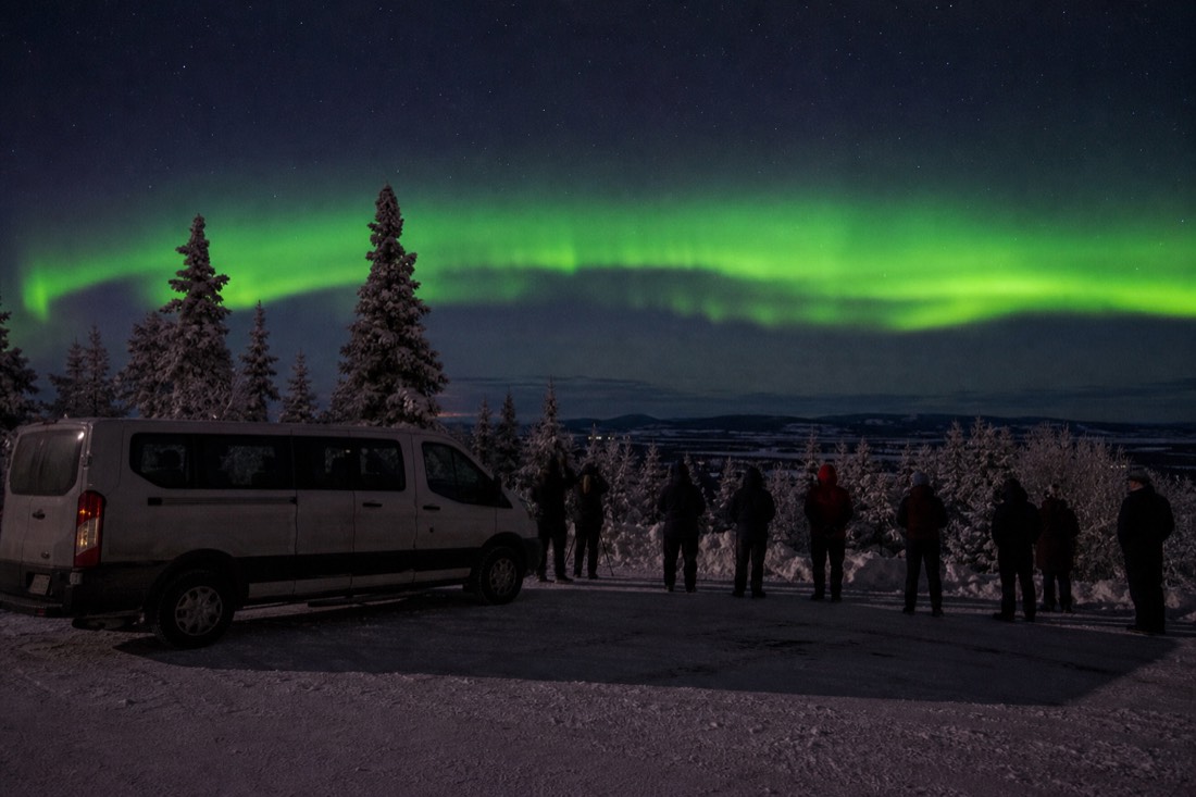 Small tour van parked at a dark roadside viewpoint while guests watch a bright aurora band over snowy trees