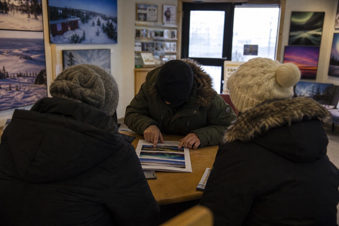 Guide in a heavy parka talking through a printed multi-night itinerary with two visitors inside a warm Yellowknife tour office