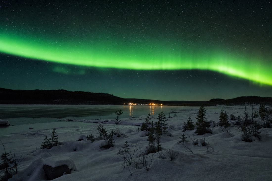 Long green aurora arc stretching across the entire night sky above tiny distant cabin lights