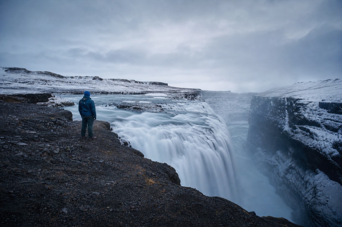 Traveler in a warm coat watching a powerful waterfall under moody grey coastal skies
