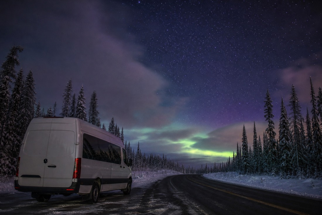 Tour van parked on a dark winter road under a sky clearing to reveal stars and faint aurora