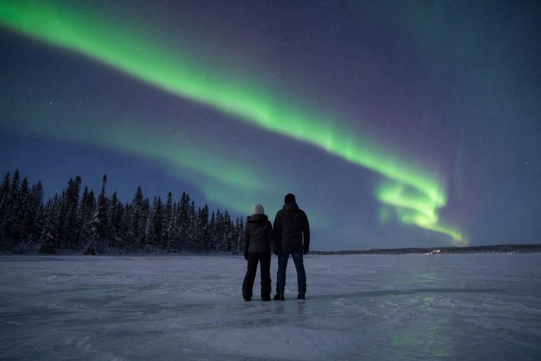 Couple standing on a frozen lake looking up at a bright green aurora ribbon overhead