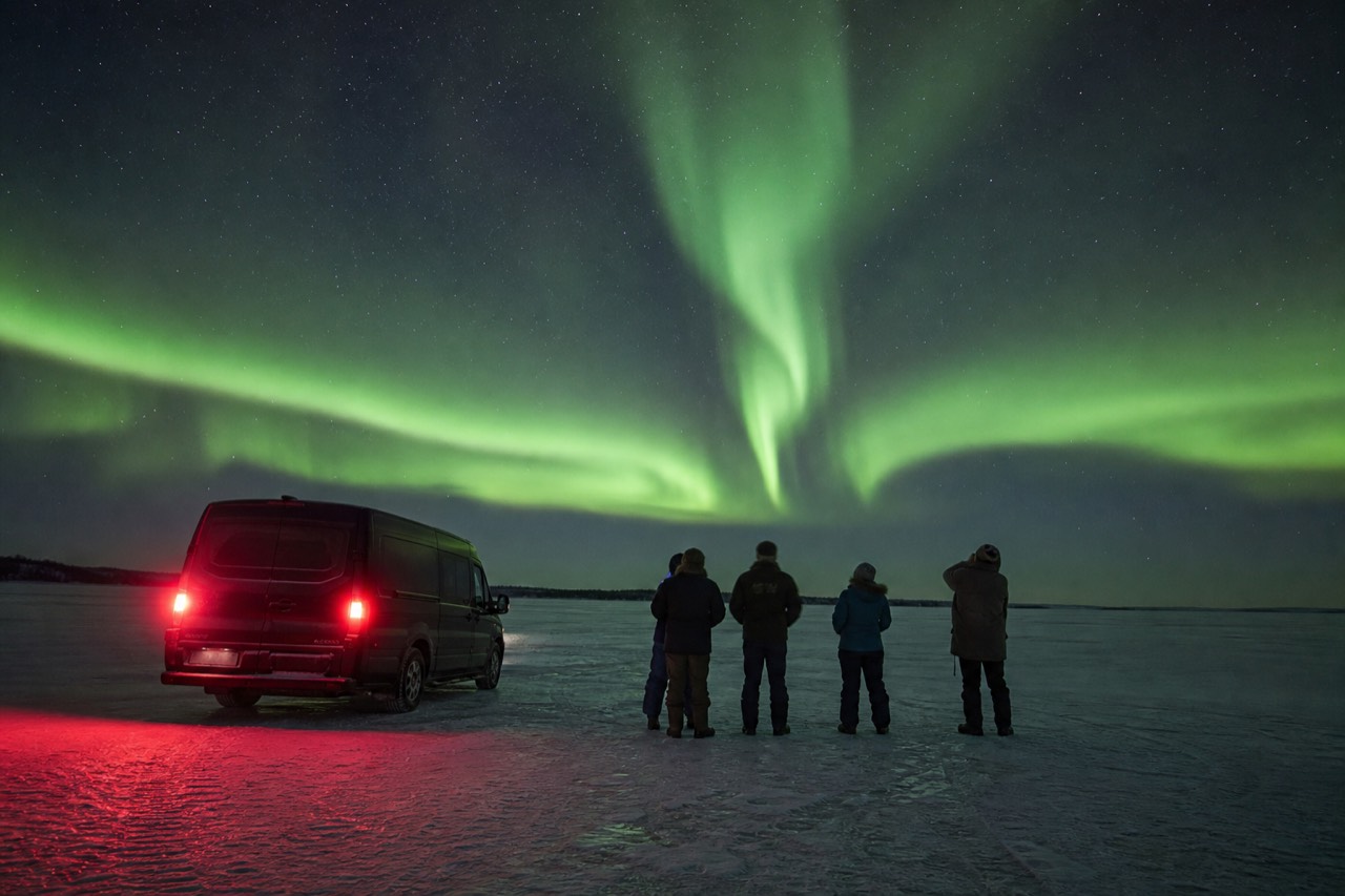 A small tour group beside a heated van watching bright green aurora over a dark frozen lake outside Yellowknife