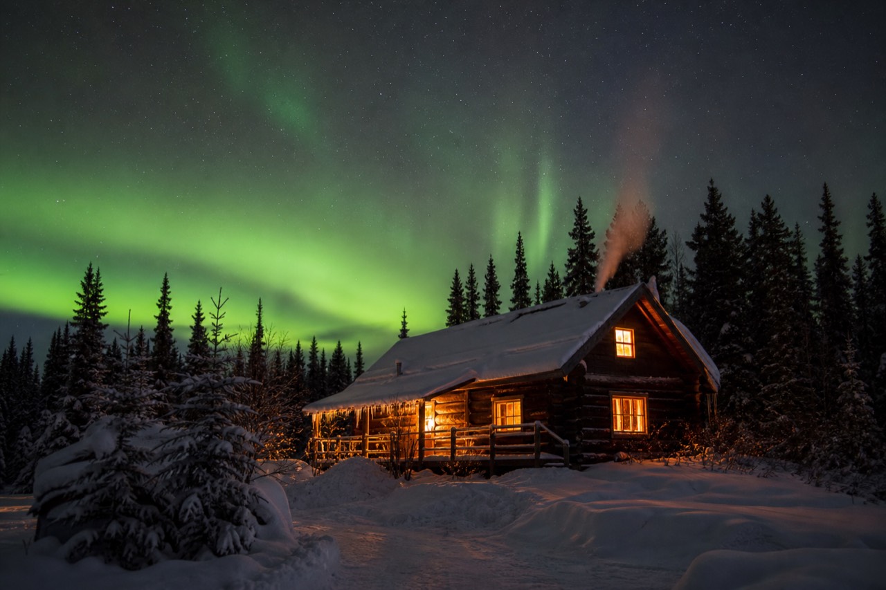 A warm log lodge glowing under a green aurora sky near Madeline Lake on the Ingraham Trail outside Yellowknife