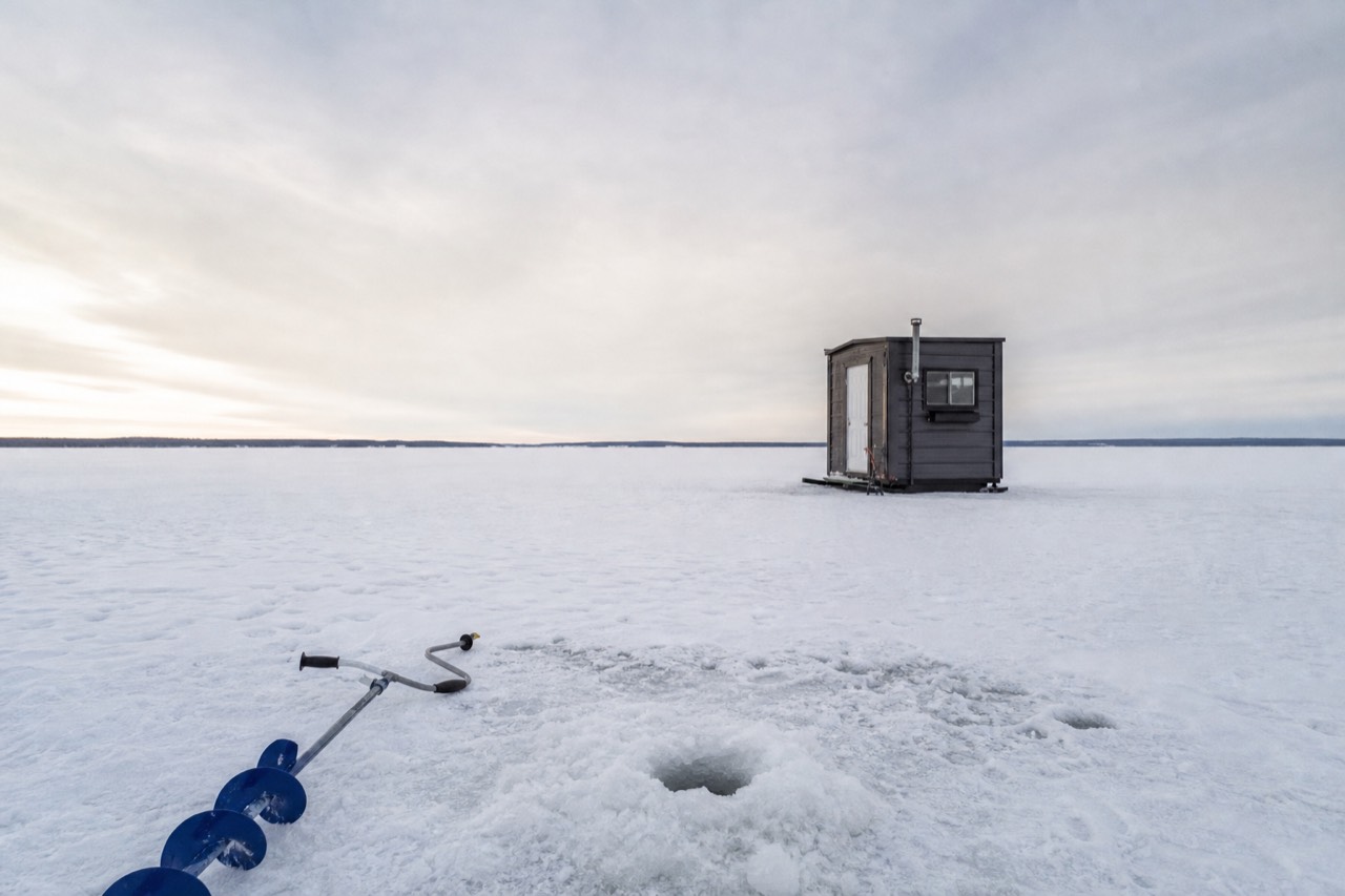 A heated ice fishing hut on the frozen expanse of Great Slave Lake with an auger and fishing hole in the foreground